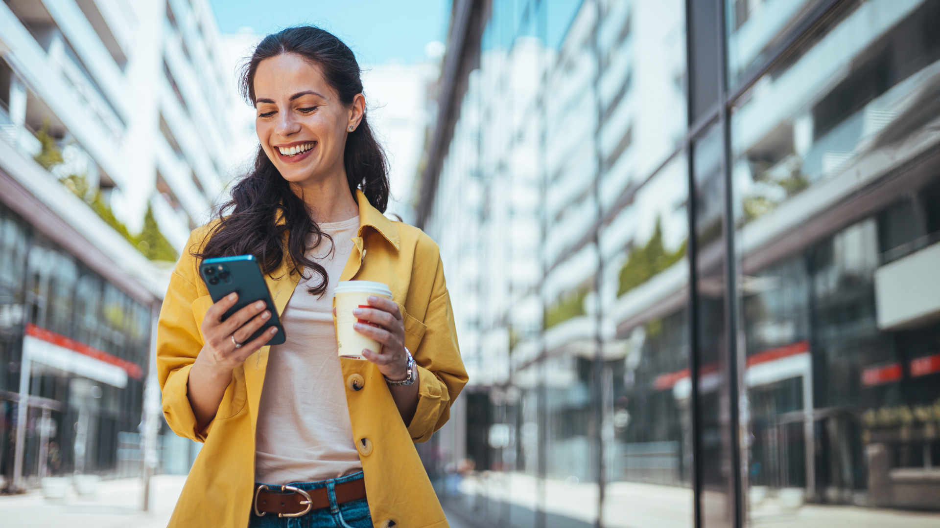 A woman looks happy whilst looking at her mobile phone, she is also carrying a paper cup of coffee