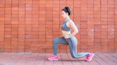 woman wearing grey crop top and leggings and pink trainers doing a lunge sideways to the camera in front of a brick wall.