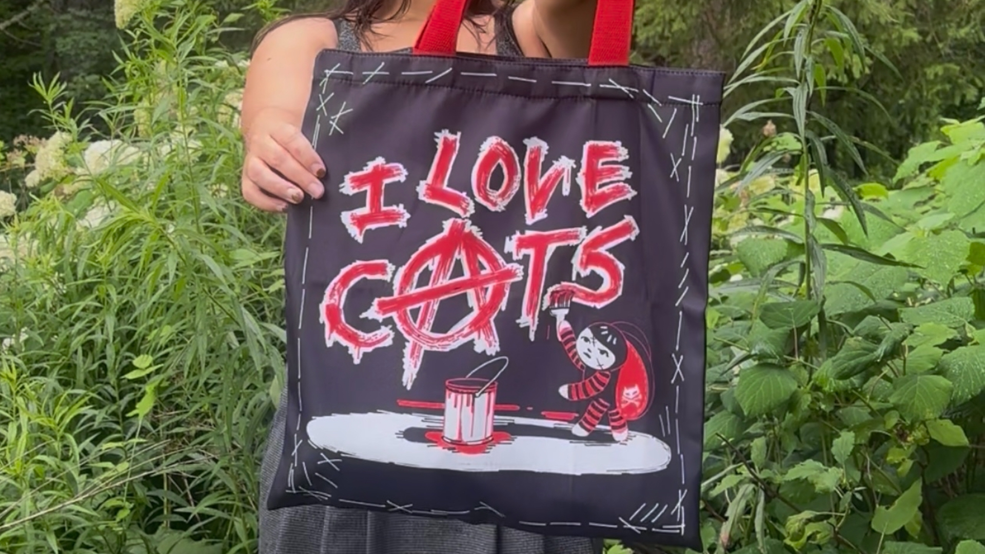A figure holds out an "I love Cats" tote bag against a green, leafy background of plants