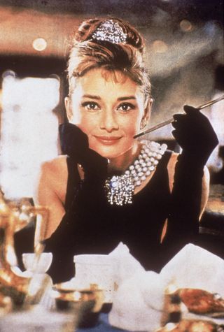 Audrey Hepburn (1929 - 1993), in a black, shoulderless dress, matching gloves, and a tiara, smiles with a cigarette holder in her hand, in her role as Holly Golightly the film, 'Breakfast at Tiffany's.'