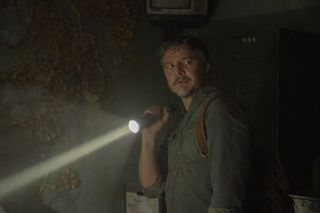 Pedro Pascal as Joel, standing in a dark room where the only light comes from a torch that he's holding. He is turned towards the camera and looking apprehensive.