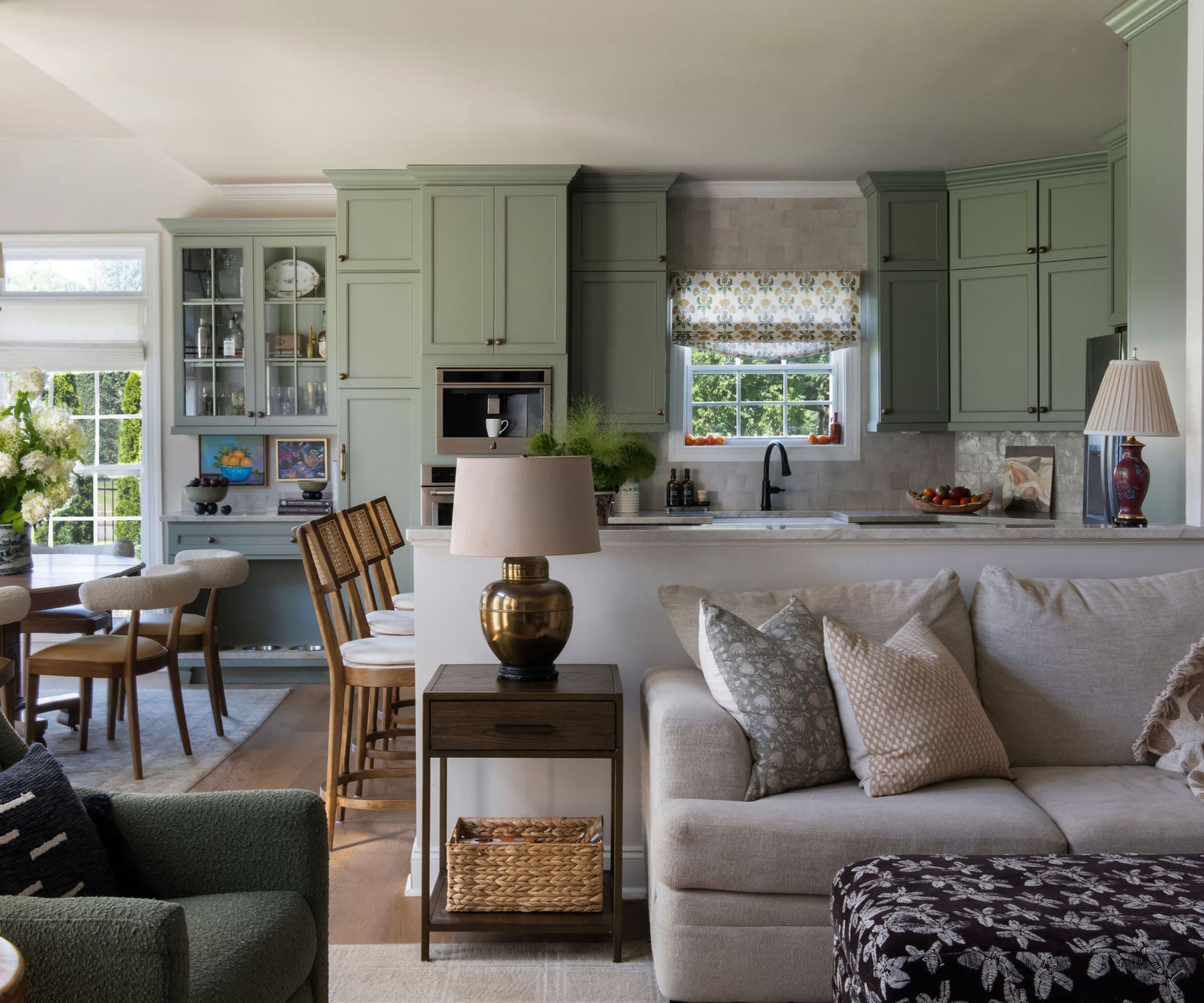 Kitchen with sage green painted cabinetry, an open plan living room with a cream sofa and block print floral pillows and ottoman, and a dining room with wooden furniture onlooking a garden.