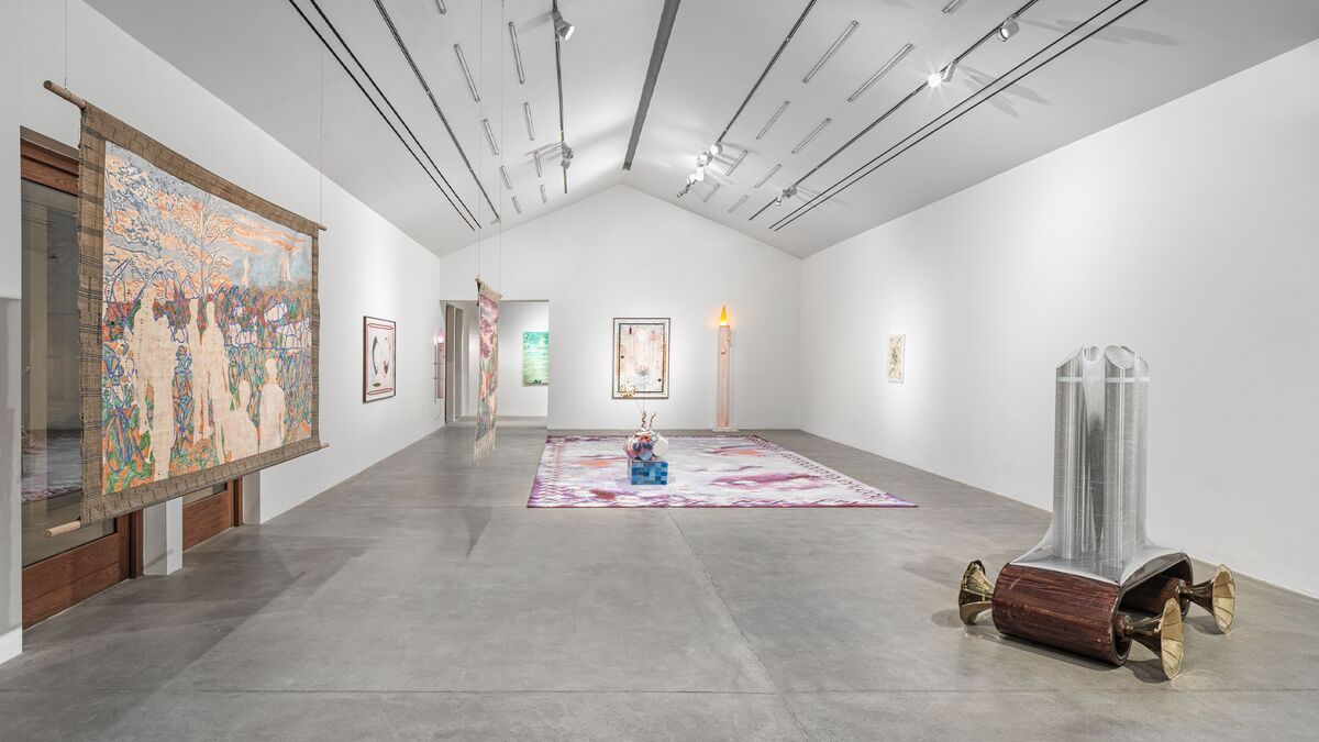 An installation view of a gallery show features a tapestry depicting multiple people in a park (on the left), a central pink and white rug, and a prototype of a car in wood (on the right).