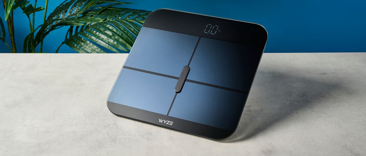 a sleek, clean-looking smart scale with tempered glass finish and a cross-shaped design on top of the glass, almost blue in the studio lighting, rests upon a table