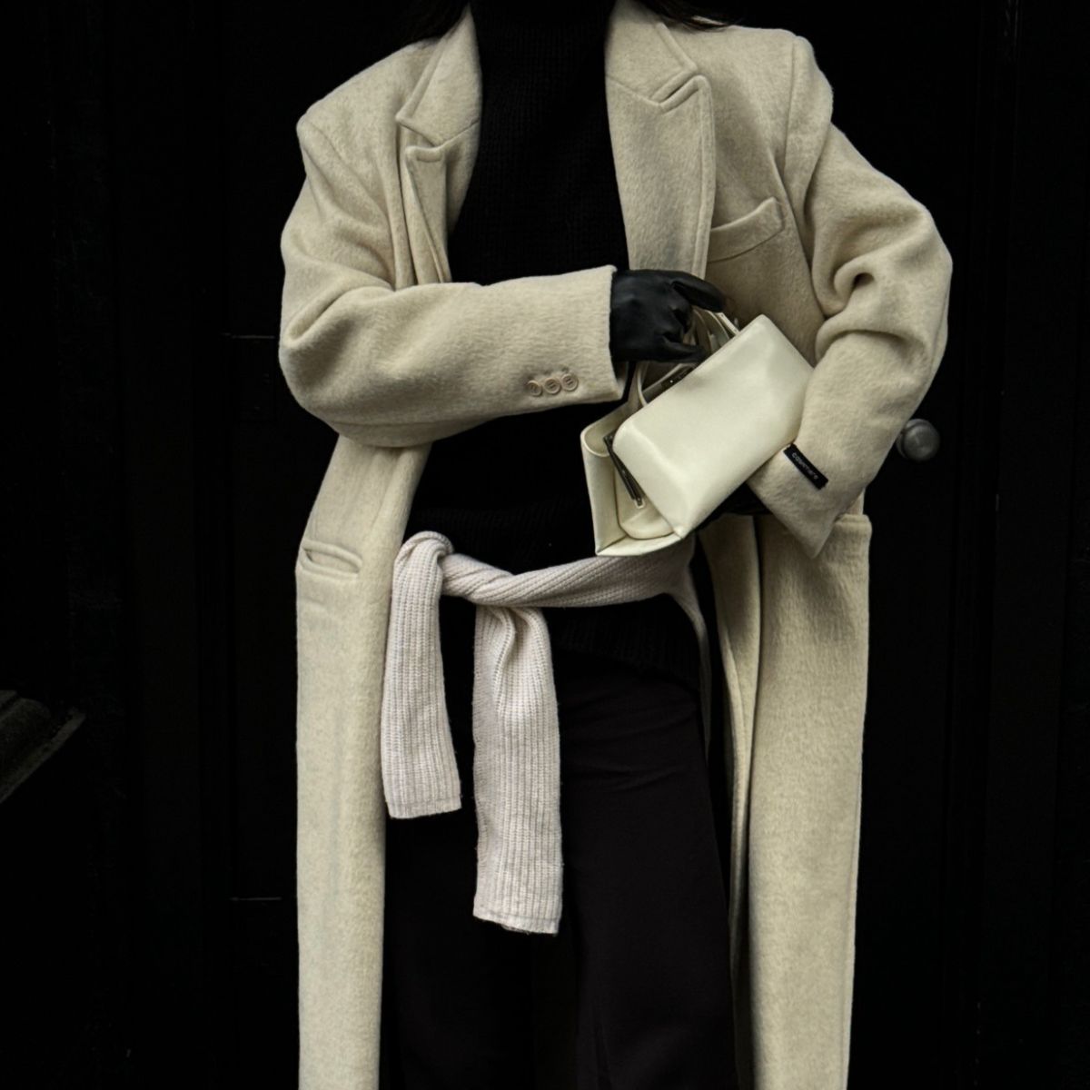 Person wears black turtleneck, cream long coat, cream handbag, black leather gloves