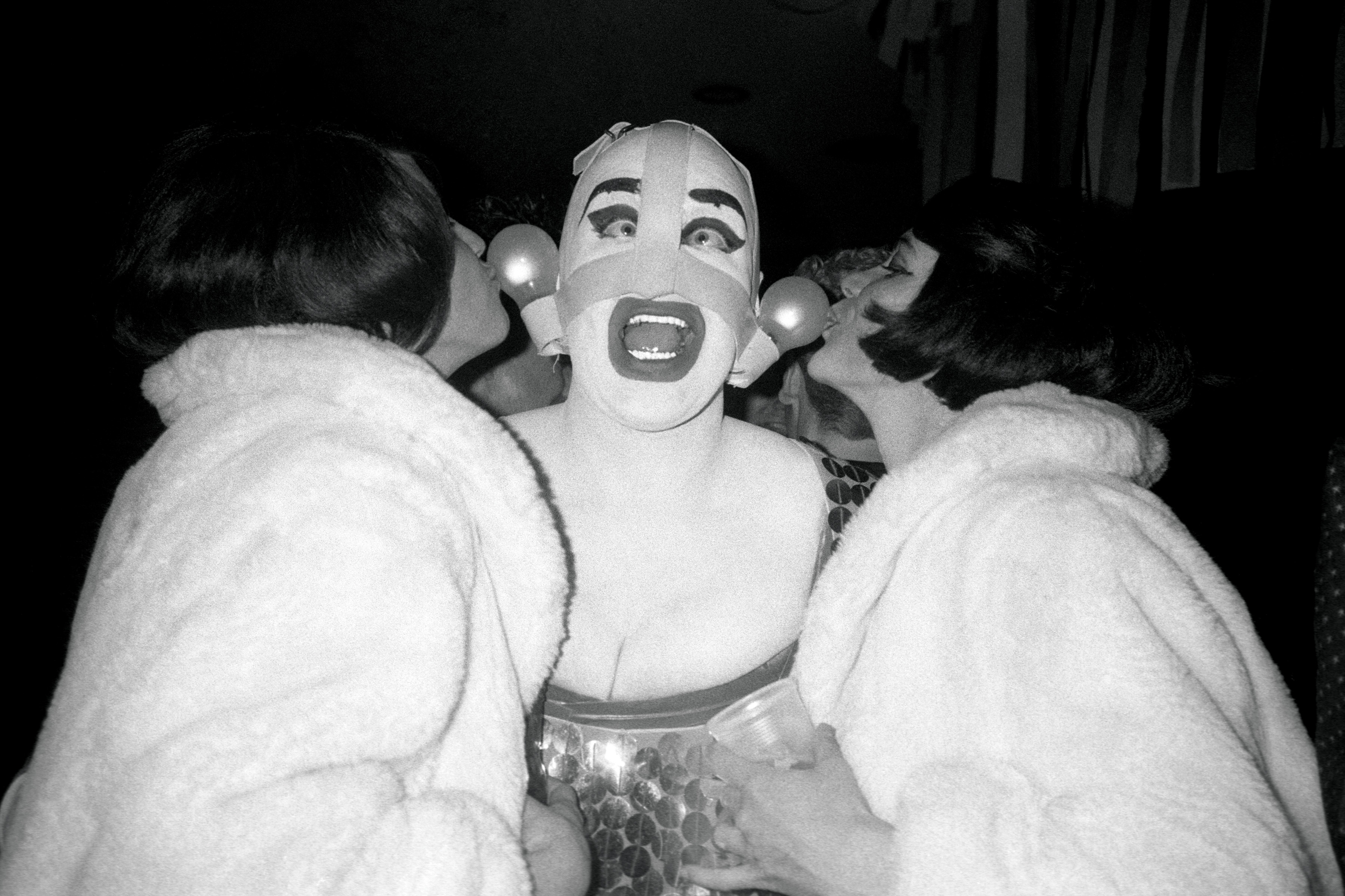 Leigh Bowery in heavy make-up and costume being kissed by two women at a party in 1988