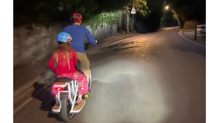 Mycle cargo Electric bike is show from the rear with a girl on the back with an adult riding the bike with lights on in the dark up a hill on the road