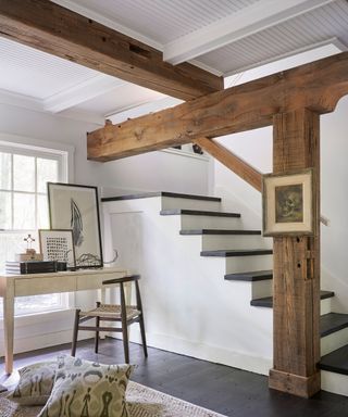 Bright white living space with warming wooden beams and pillars, white paneled wooden ceiling, black flooring, wooden desk and chair beside window, rug and cushions on floor