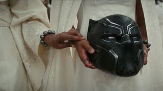 Still from the movie Black Panther Wakanda Forever (2022). Here we see a close up of a women wearing white robes who is holding the Black Panther helmet (black cat-like helmet with silver detailing) in her hands. Another woman wearing white robes next to her is reaching out and holding onto the helmet holders hand.