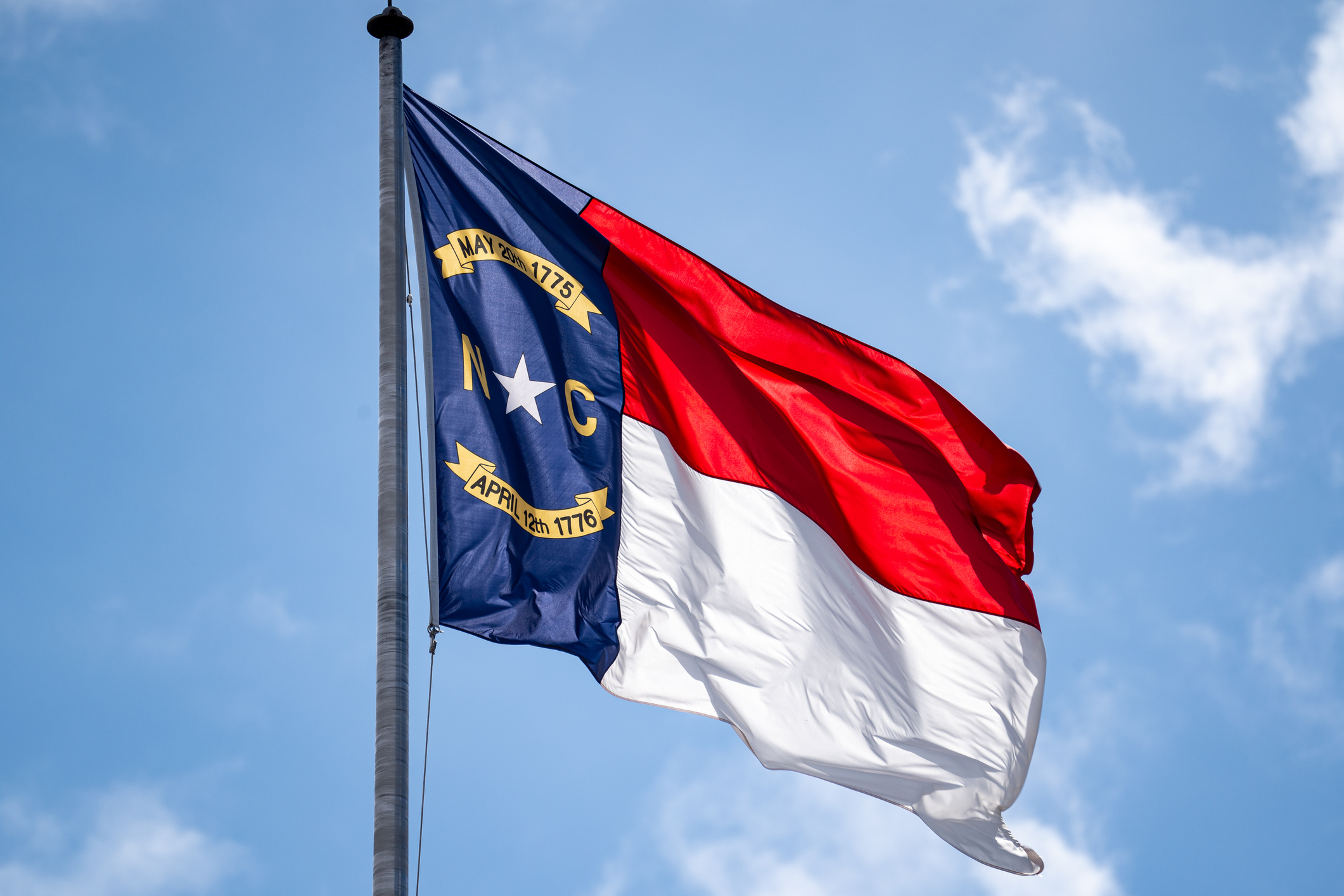Close-up photo of a North Carolina state flag waving in the wind against a blue sky
