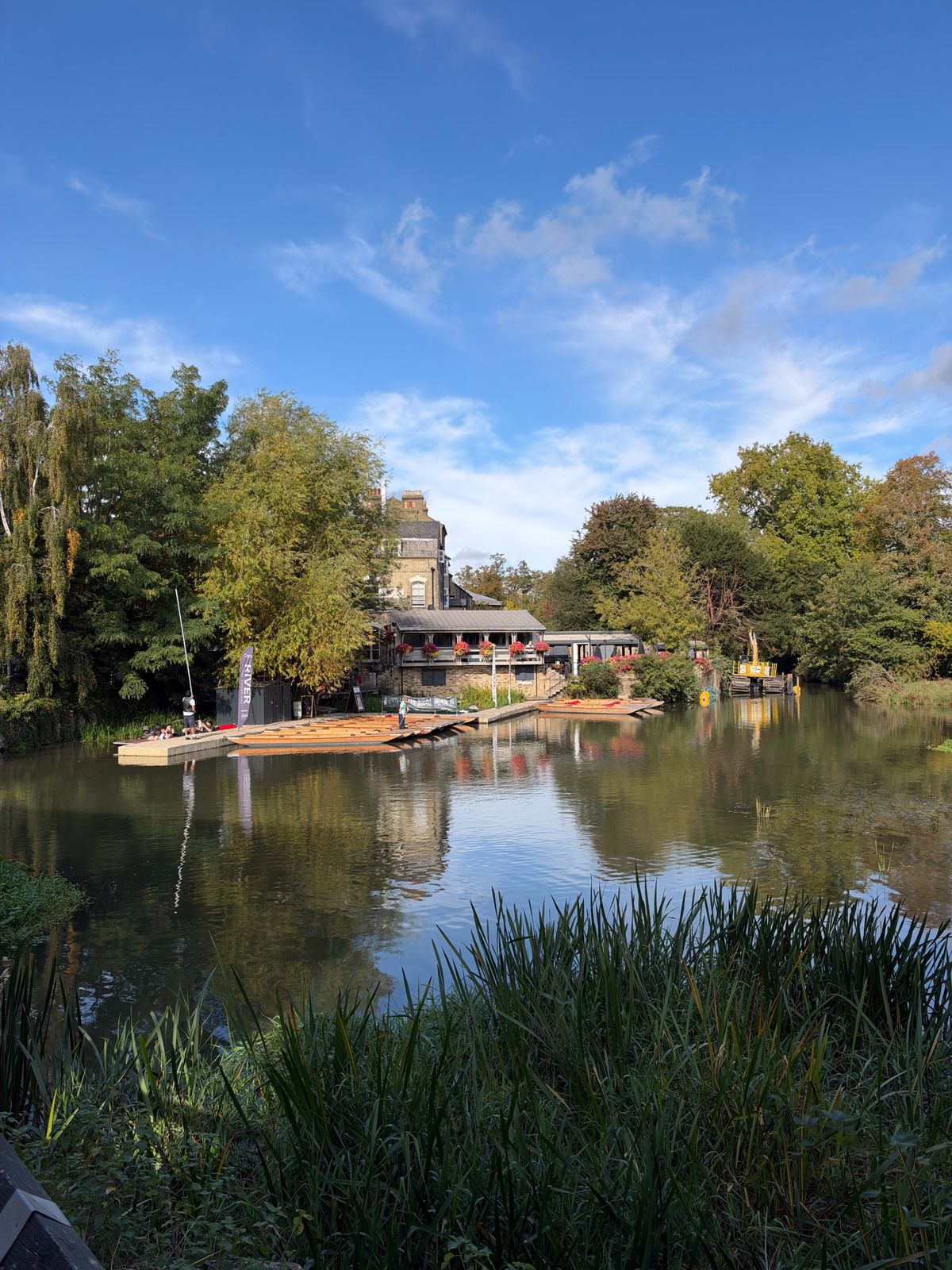 Calm river bordered by reeds and trees, with wooden punts and a riverside building decorated with hanging flowers.