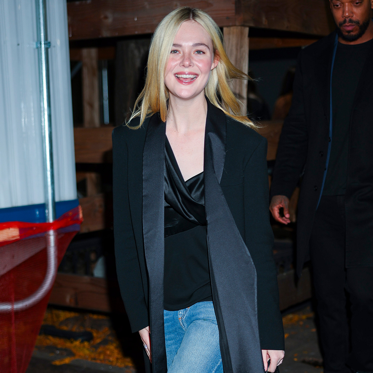 Elle fanning wears a long black coat, black blouse, and blue jeans.