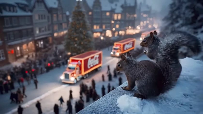 Two squirrels sit on a snowy ledge overlooking a town square at night, where two illuminated Coca-Cola Christmas trucks drive through a crowd of people near a decorated Christmas tree.