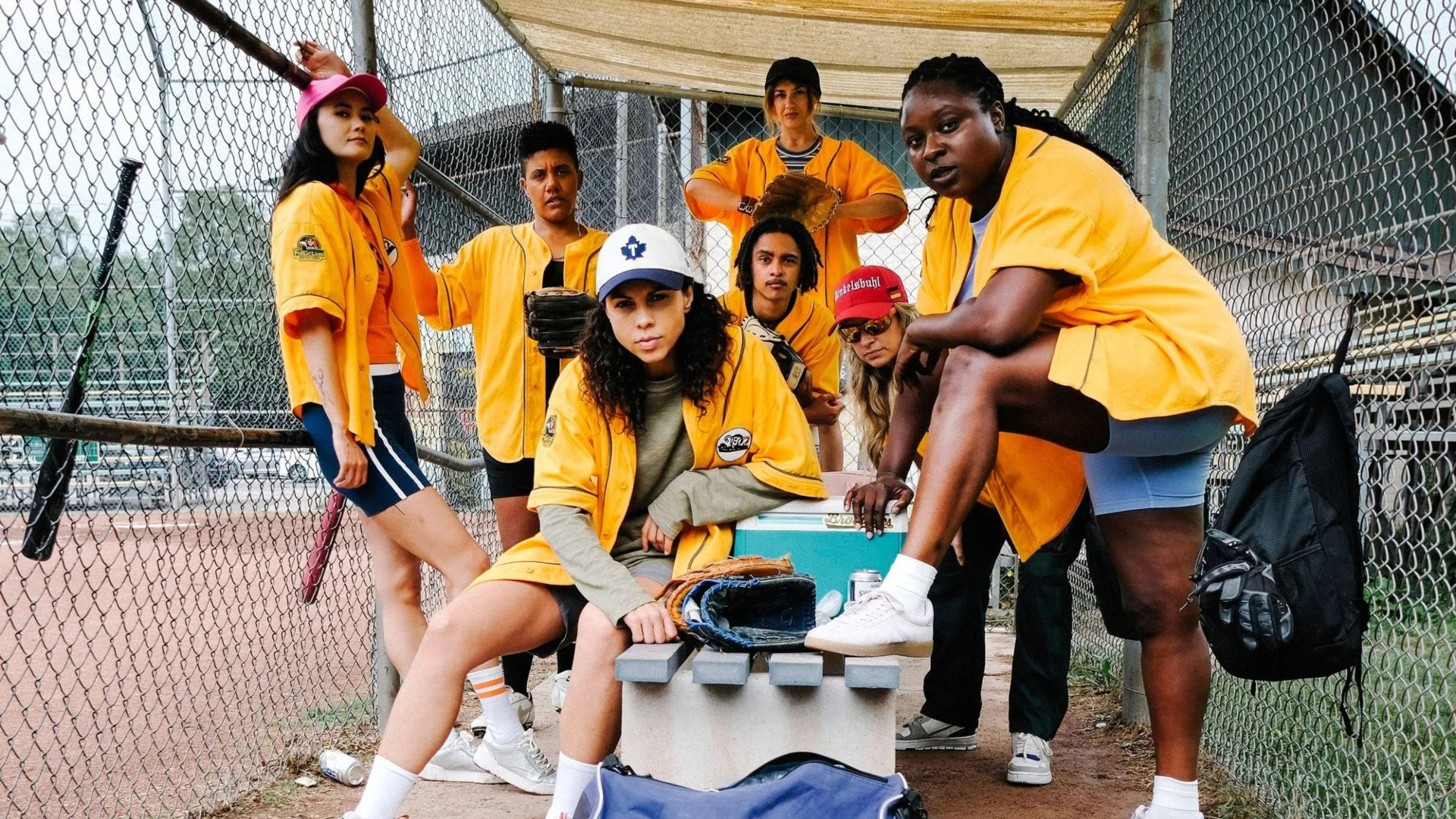 A group of women wear yellow jerseys while posing in a baseball dugout, in the TV series 'Slo Pitch.'