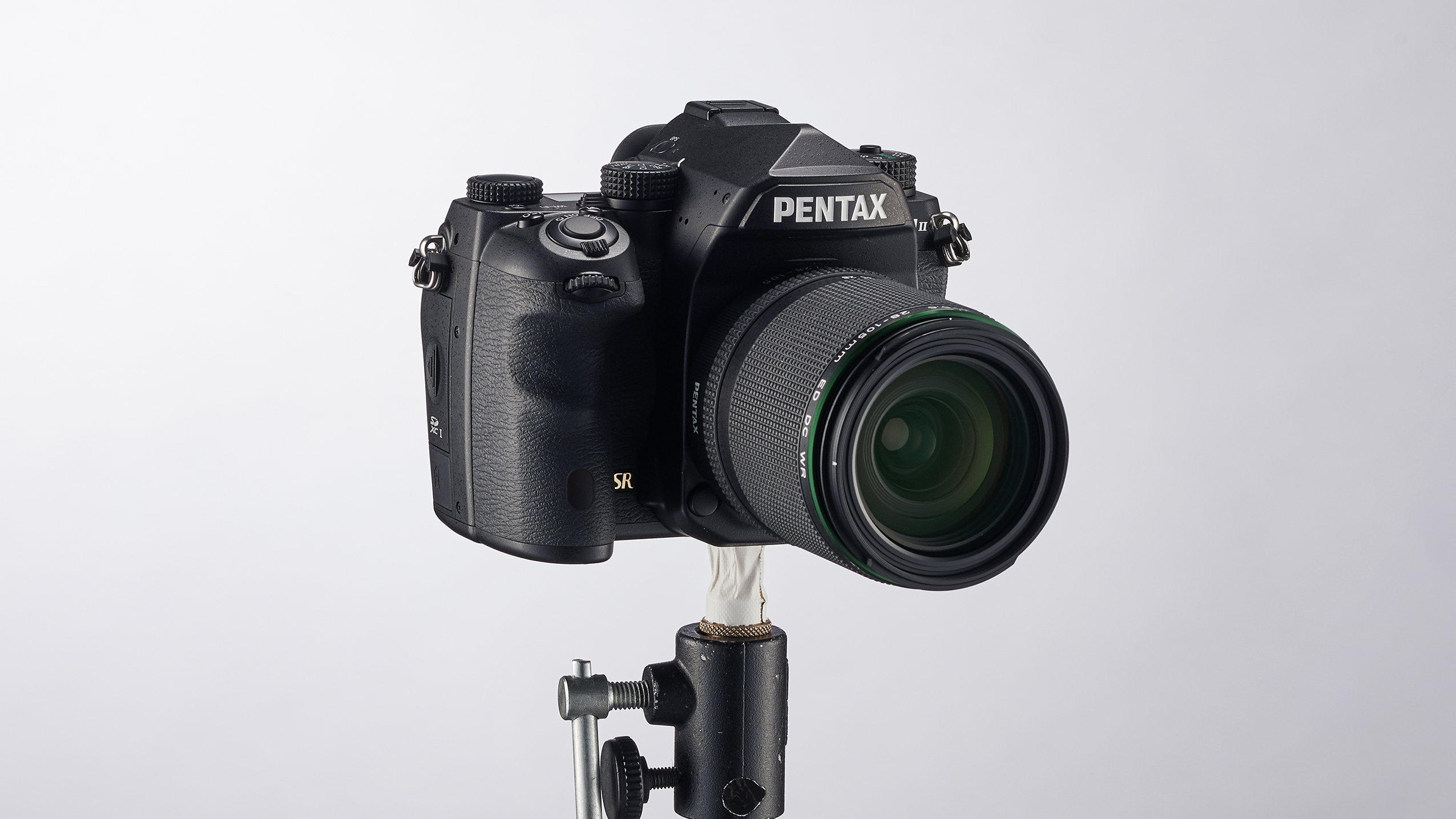 Black Pentax K-1 Mark II DSLR camera with zoom lens mounted on a small metal clamp against a plain white background