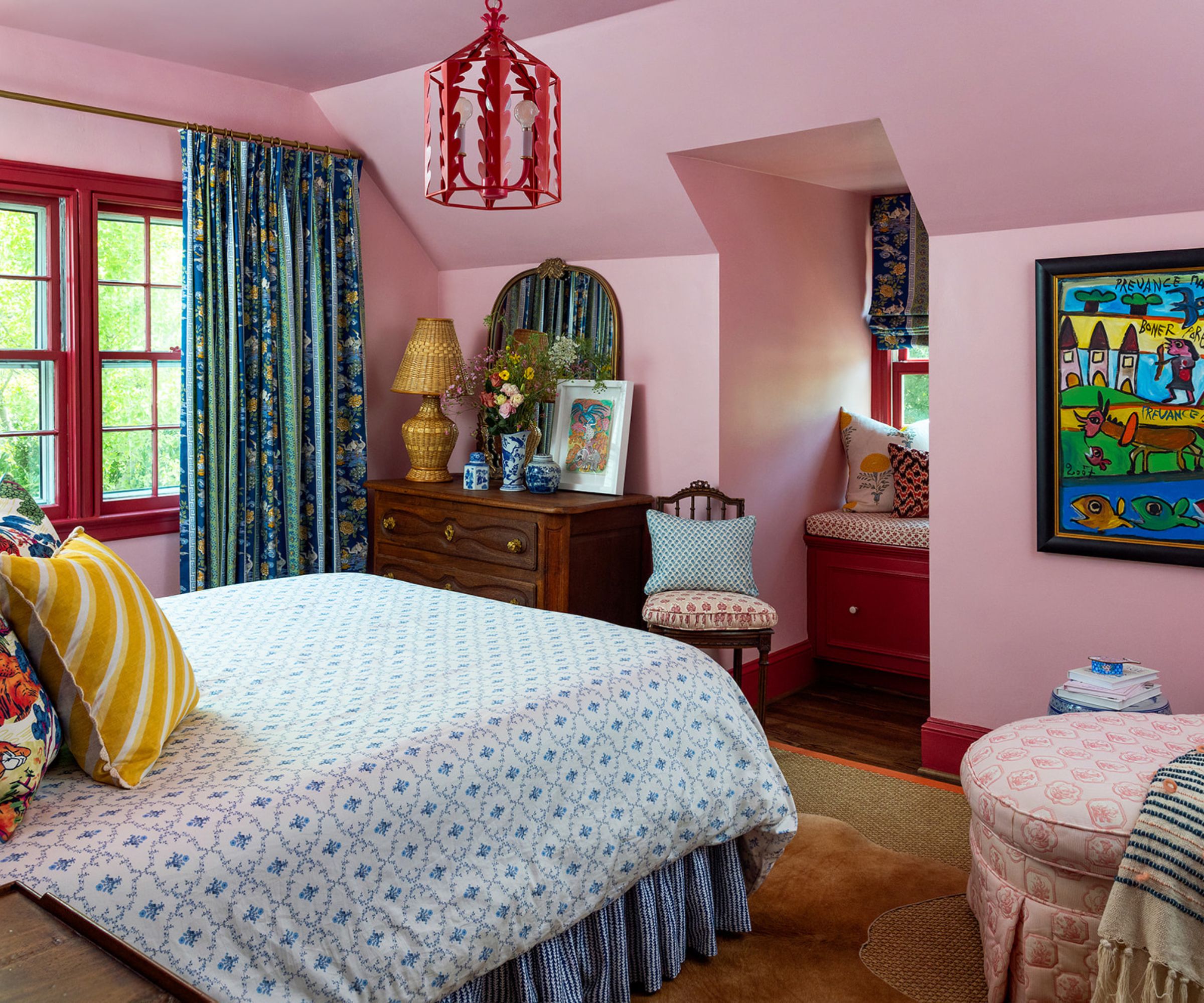 A cozy and eclectic bedroom with mid-tone pink walls, red window trim, a bed with patterned blue bedding, a wooden dresser, a bright red pendant ceiling light, and blue, green, and yellow artwork.