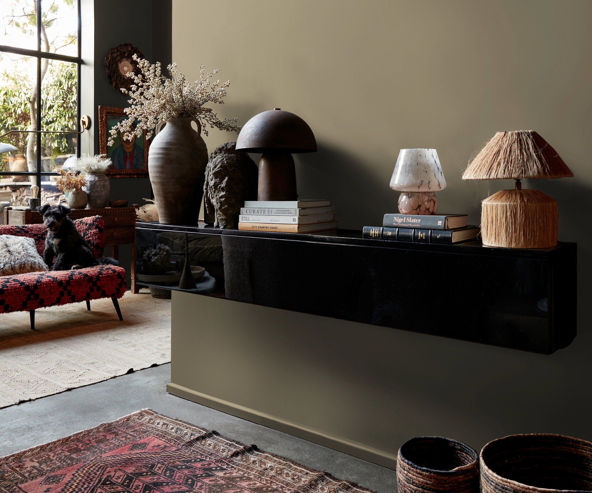 Black glossy console with lamps, vases, books, and a dog on a red patterned sofa in a moody, eclectic room.