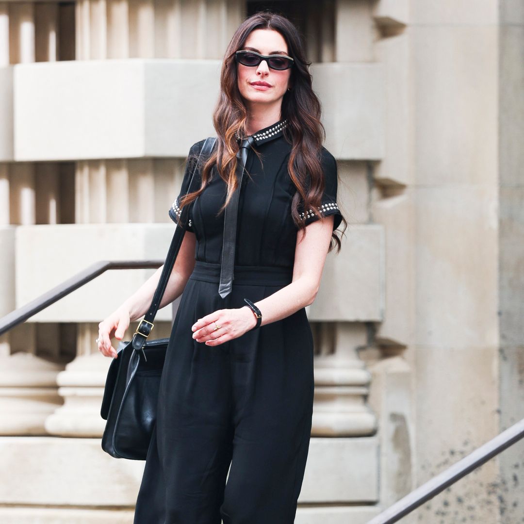 Anne Hathaway wearing a black collared shirt, tie, messenger bag, and sunglasses. She has curls in her brown hair. 