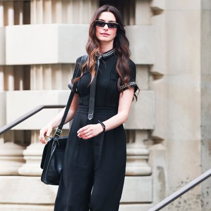 Anne Hathaway wearing a black collared shirt, tie, messenger bag, and sunglasses. She has curls in her brown hair.