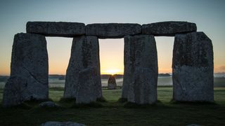 four pillars of stonehenge are visible at the front of the image. Behind is the heel stone with the sun touching the top of it. far in the back are trees and fog