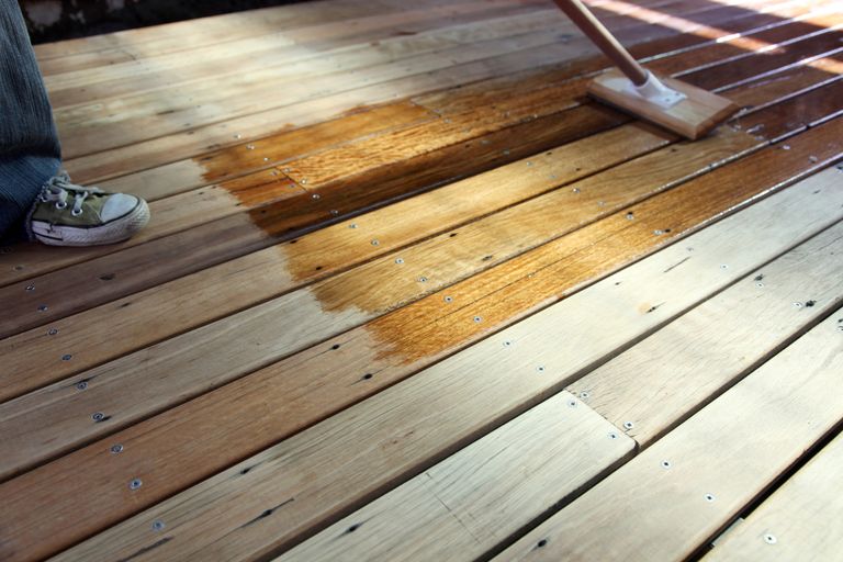 A person wearing denim jeans and casual green and white laced sneakers applying decking oil to wooden decking with someone applying decking oil to wooden deck with long-handled wooden brush