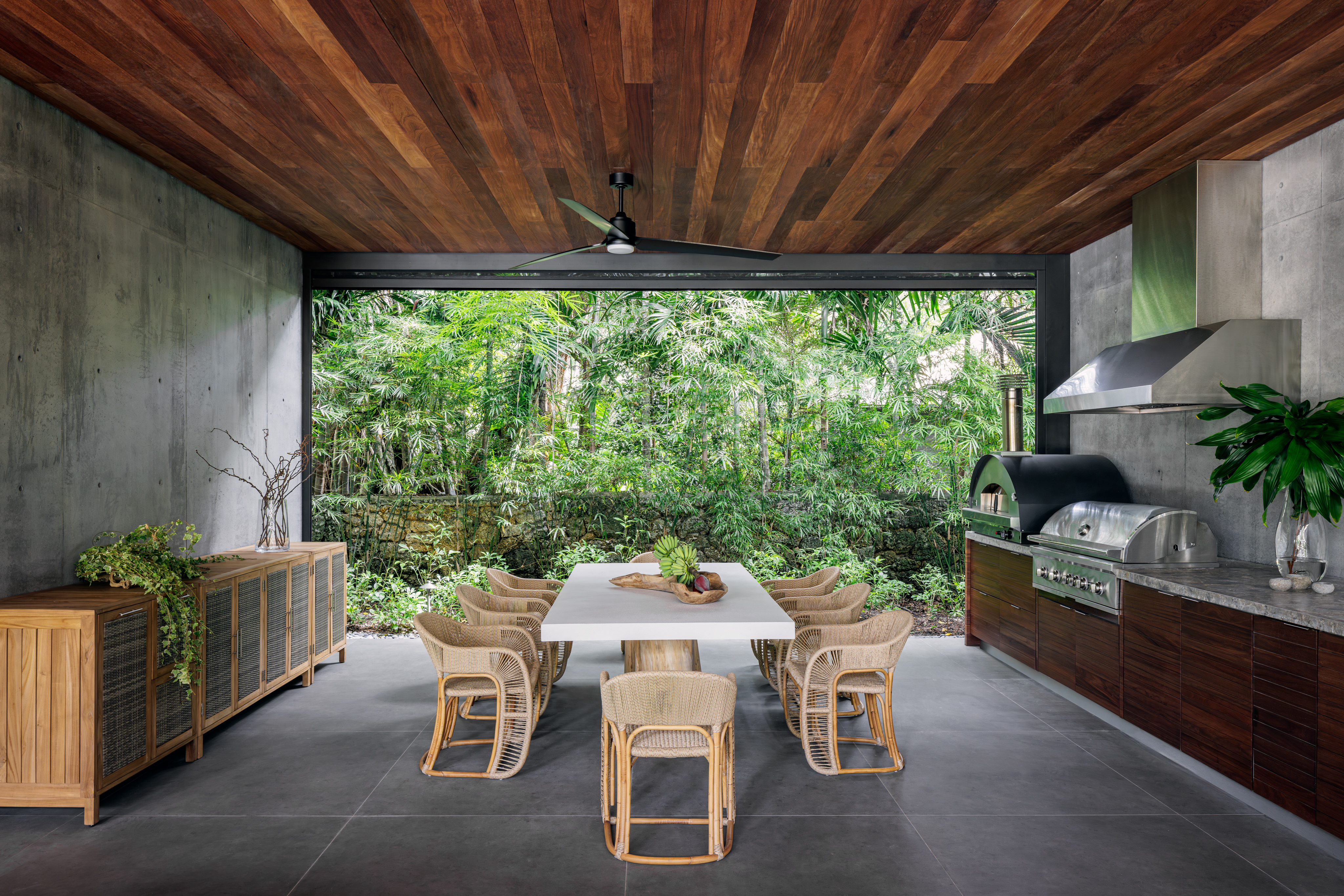 An outdoor space with a wooden roof, cement flooring, a large dining table and a sideboard with grill