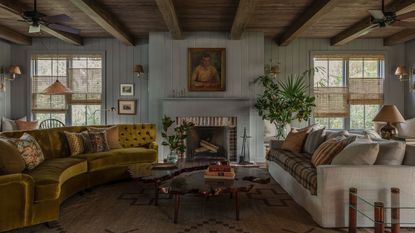 A large, light blue living room with wooden ceilings and a cozy atmosphere. There are two sofas (one white, one ochre), lots of paintings on the wall, and separating blinds on the windows.