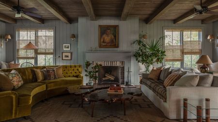 A large, light blue living room with wooden ceilings and a cozy atmosphere. There are two sofas (one white, one ochre), lots of paintings on the wall, and separating blinds on the windows.