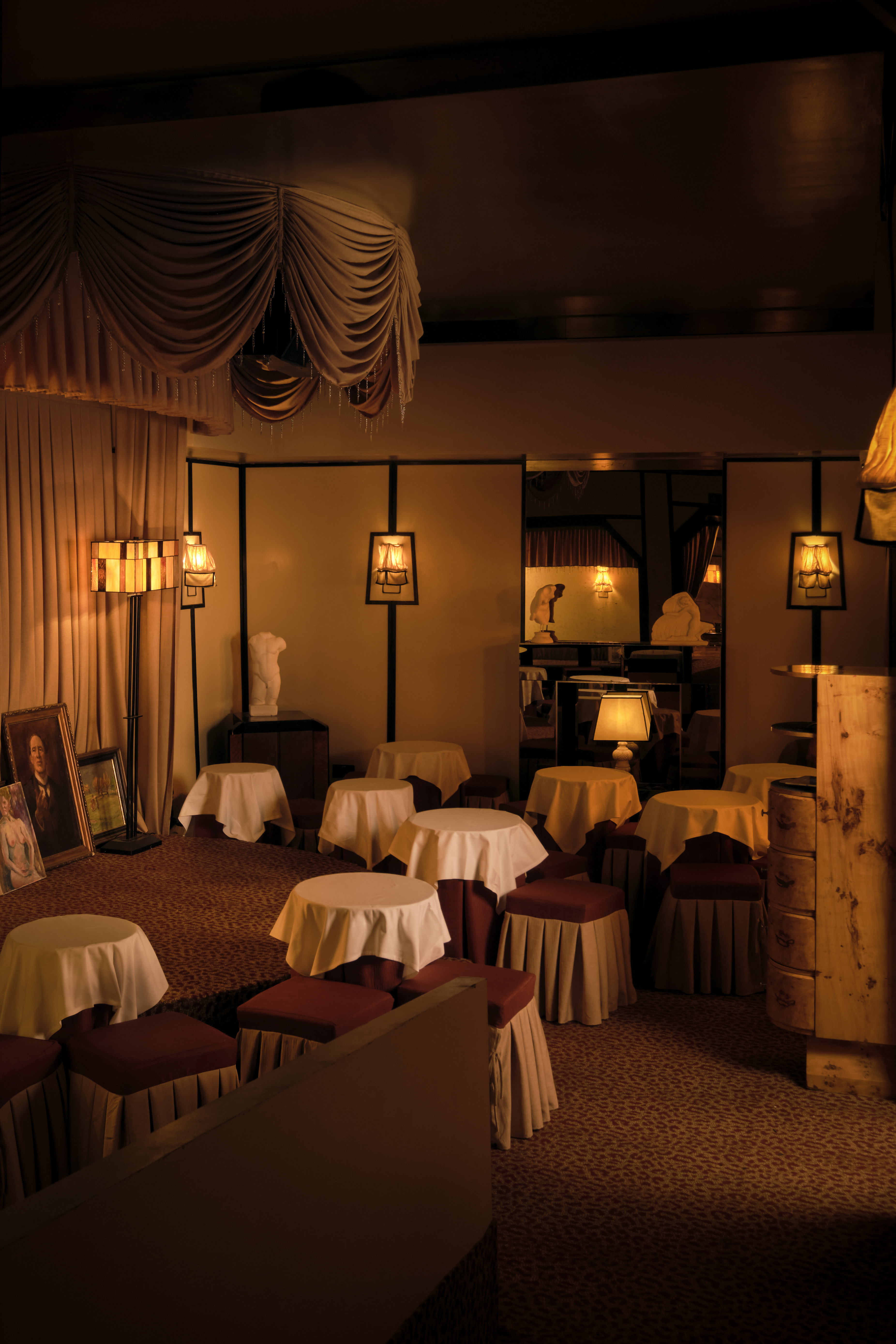 An amber-lit nightclub features retro-inspired furniture echoing the atmosphere of the 20th century, marble busts, plush fabrics, and wall murals.