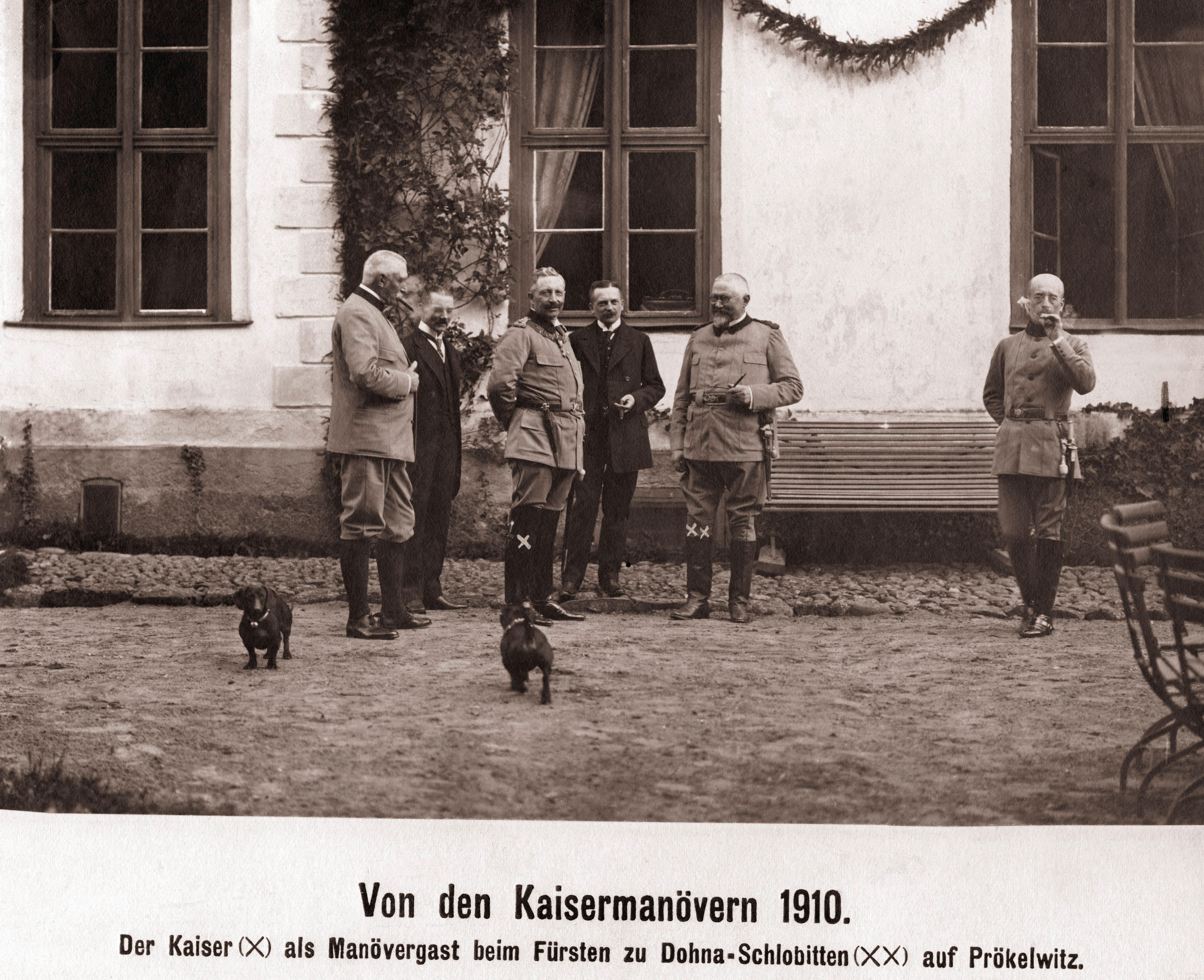 A sepia-toned photograph from 1910 showing Emperor William II standing with military officers and huntsmen in a cobbled courtyard outside a country house, with two dachshunds in the foreground. The men wear hunting uniforms, and the dogs stand alert near their feet.