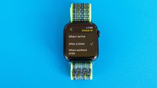 Detail shot of an Apple Watch SE on a bright blue background showing the steps to set up and send a Check In notification in watchOS 11 