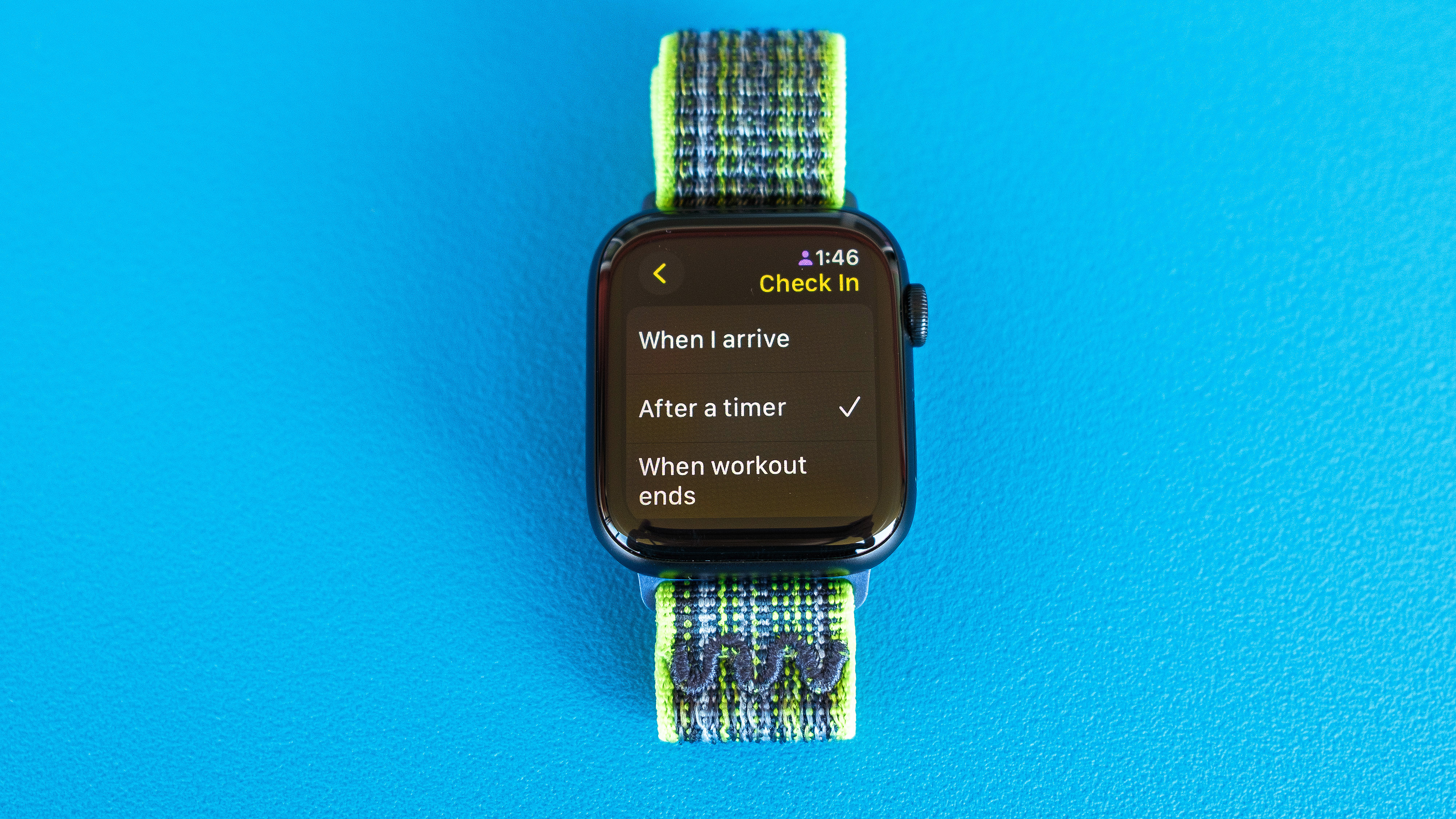 Gambar detail Apple Watch SE pada latar belakang biru cerah yang menunjukkan langkah-langkah untuk menyiapkan dan mengirim pemberitahuan Check In di watchOS 11 