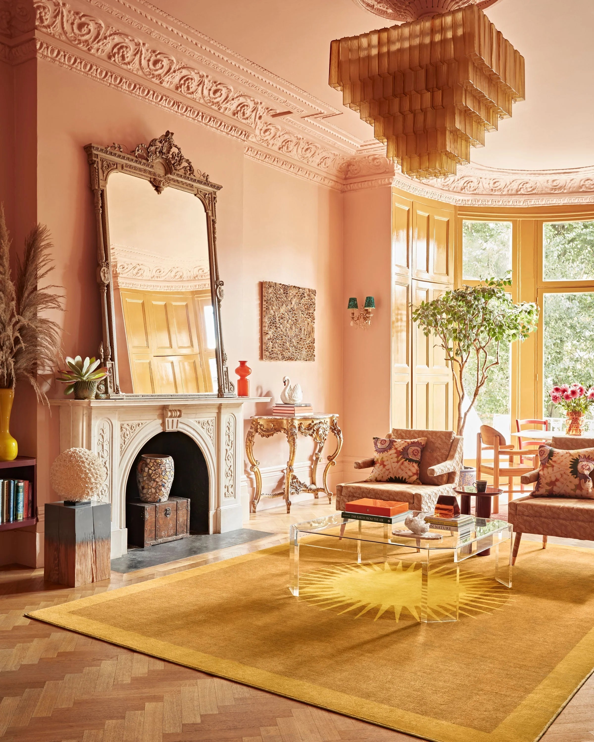 A sunshine-tinted room with stuccoed ceilings, pink walls, a room-wide, yellow rug with a stylized sun at its center, a murano glass chandelier in an amber tone, plants, and plenty of decorative objects.