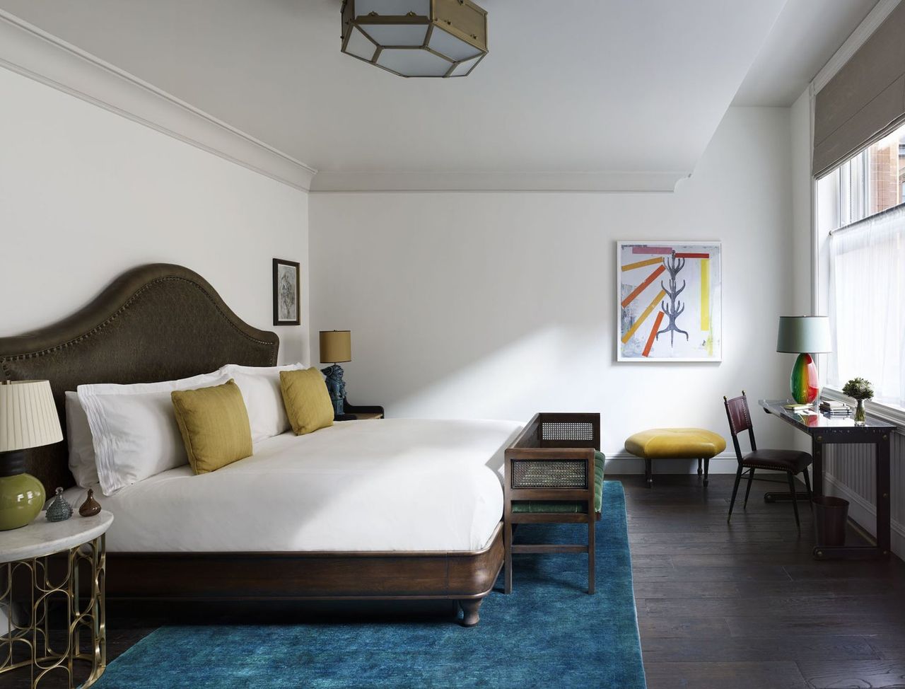 A transitional luxury hotel room features dark wood furniture including a king bed, an upholstered bench, and a chair and table as well as pastel-shaded art and accessories.
