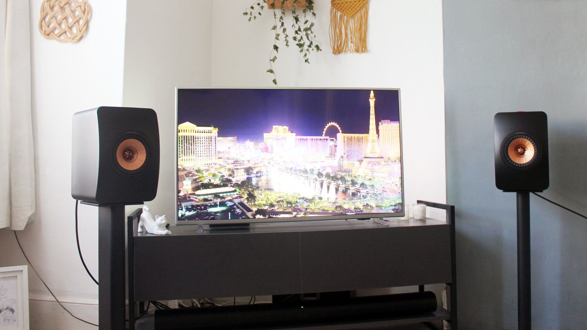 Two Kef LS50 speakers sit in a living room, on speaker stands. They are on either side of a TV, which has an image of the Las Vegas skyline on it.