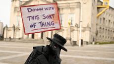 A protester dressed as a plague doctor carrying a placard saying 'down with this sort of thing'