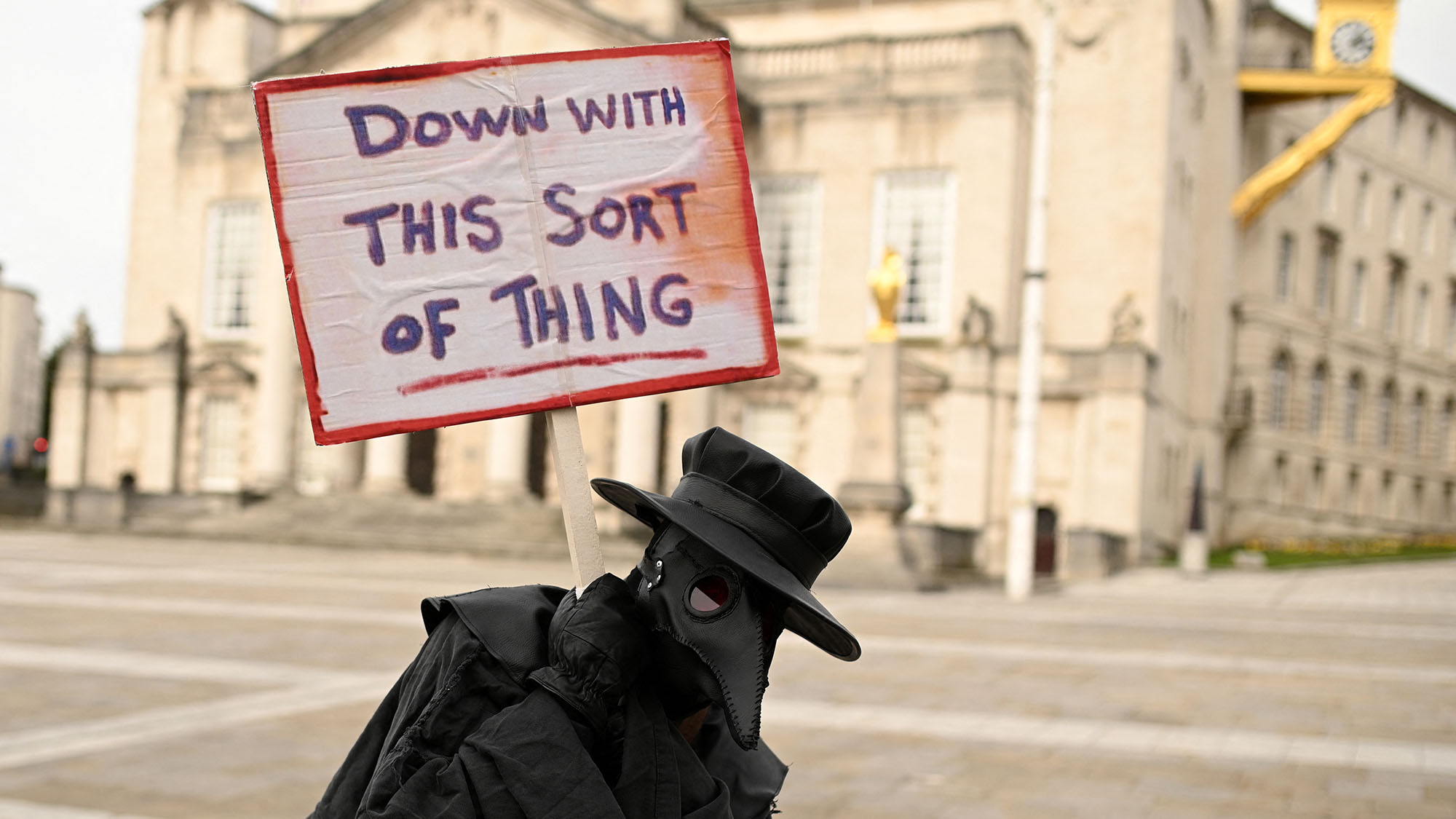 A protester dressed as a plague doctor carrying a placard saying 'down with this sort of thing'