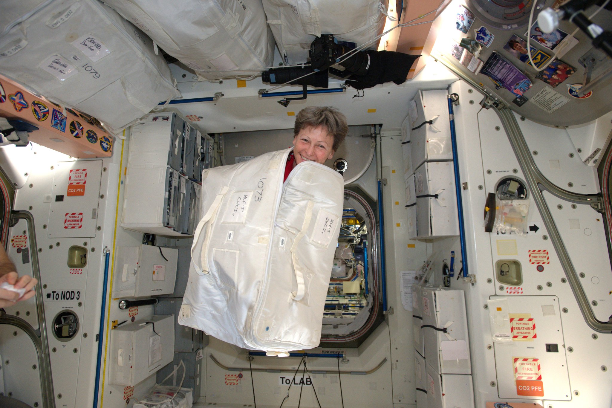 Peggy Whitson is smiling as her head pops out of the partially unzipped white cargo bag while 'floating' in zero gravity on the International Space Station.