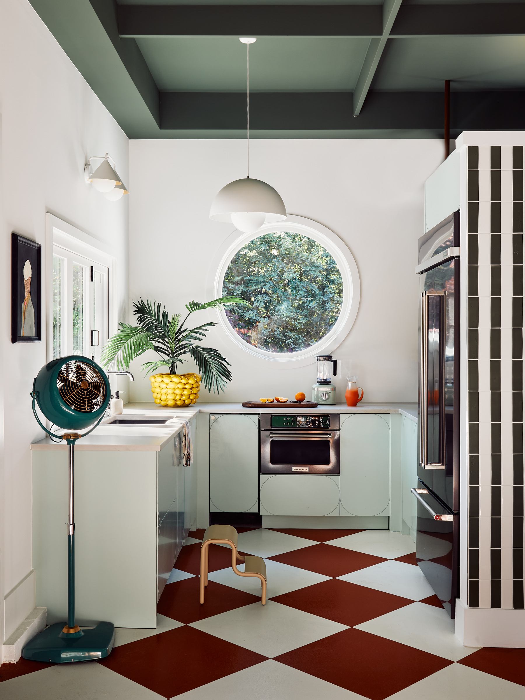 A retro kitchen with a red and white checkerboard floor, white walls, white pendant light, and circular window.