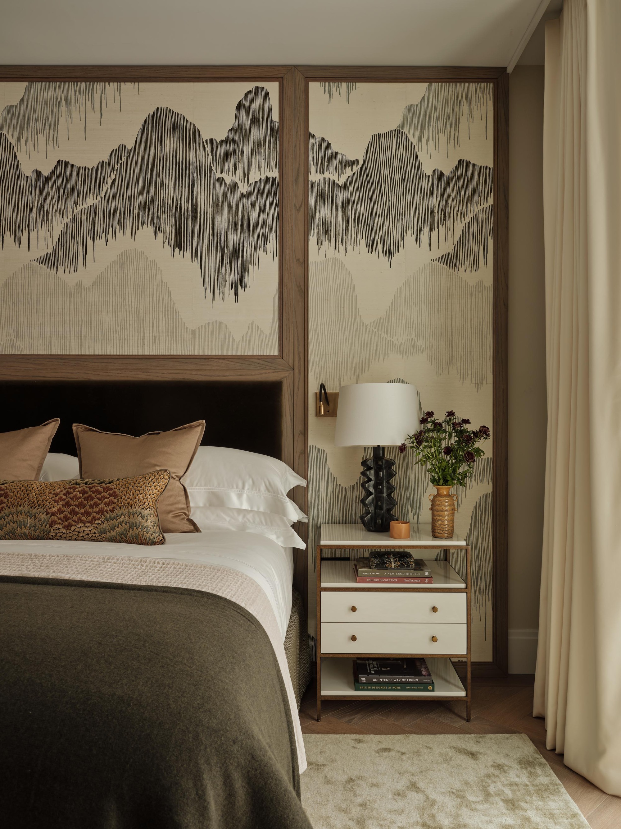 A bedroom with patterned wallpaper, a bed styled with beige cushions and a floral bolster pillow, and a styled nightstand