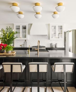 Kitchen with white painted walls, black kitchen cabinets and countertops, six white and metallic large ceiling lights above island, black metal counter chairs with upholstered seat and back cushions, tray styled with flowers and plants on island
