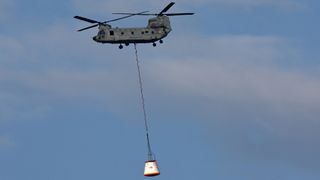 A helicopter lifts a large white crew module hoisted by a long rope in the air