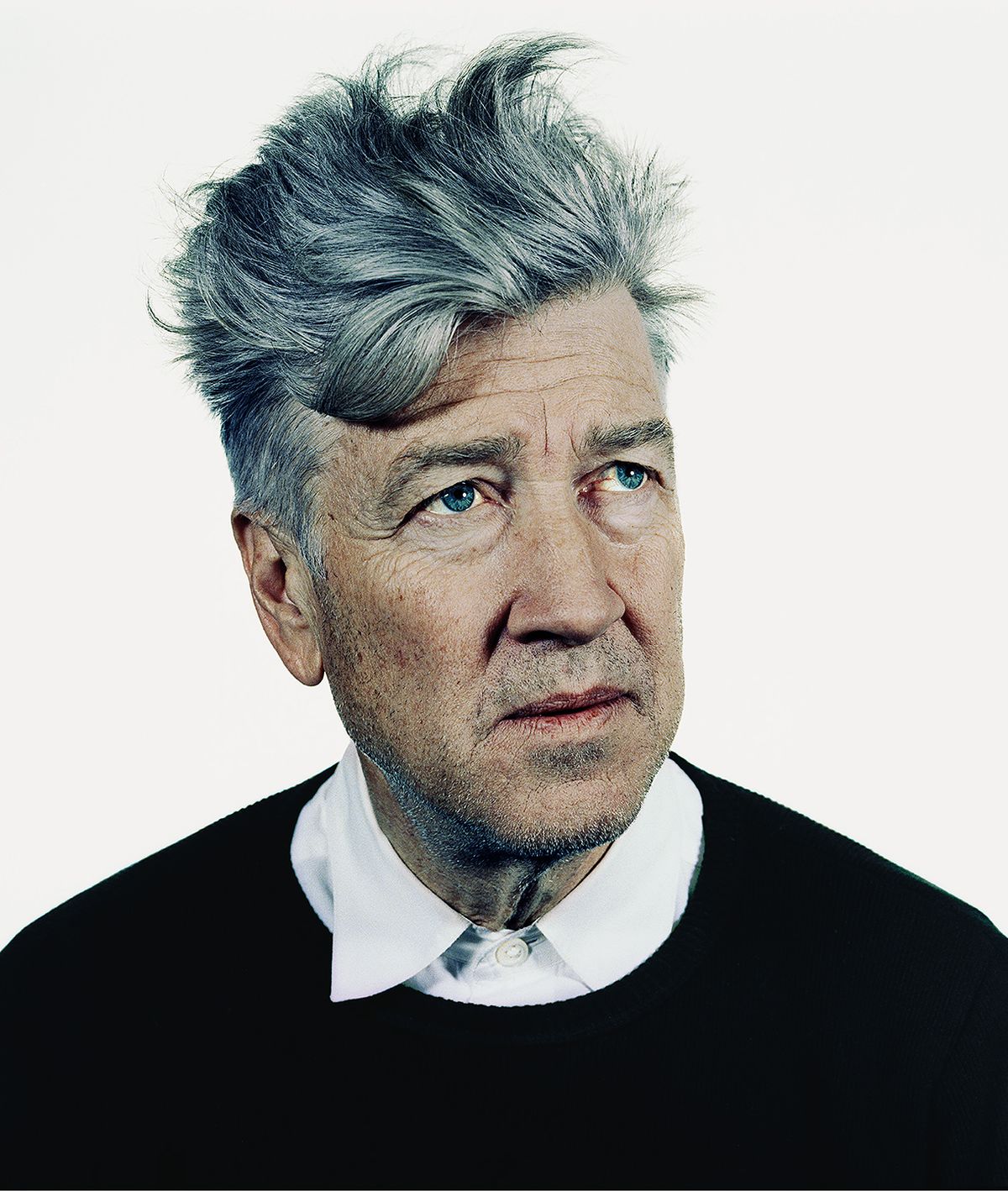 A colour portrait of David Lynch with tousled grey hair, wearing a black jumper over a white collared shirt, gazing upwards against a plain white background.
