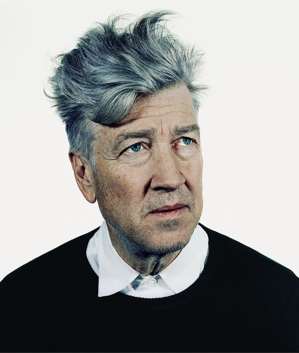 A colour portrait of David Lynch with tousled grey hair, wearing a black jumper over a white collared shirt, gazing upwards against a plain white background.