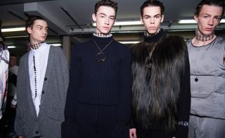 Four male models wearing looks from JW Anderson's collection. Two models are wearing blue ribbed pieces, one has a silver chain with circle pendant on and the other has a long hair fur panel covering the chest. The other two models are wearing grey pieces, one with a repeating pattern and the other has a white shirt with dark detail underneath. All models are wearing thick clear chokers with metal studs
