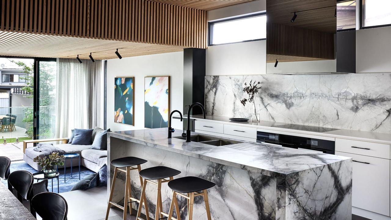 A marbled kitchen island with a backsplash of the same design. Bar stools sit by the island of an open plant space. 