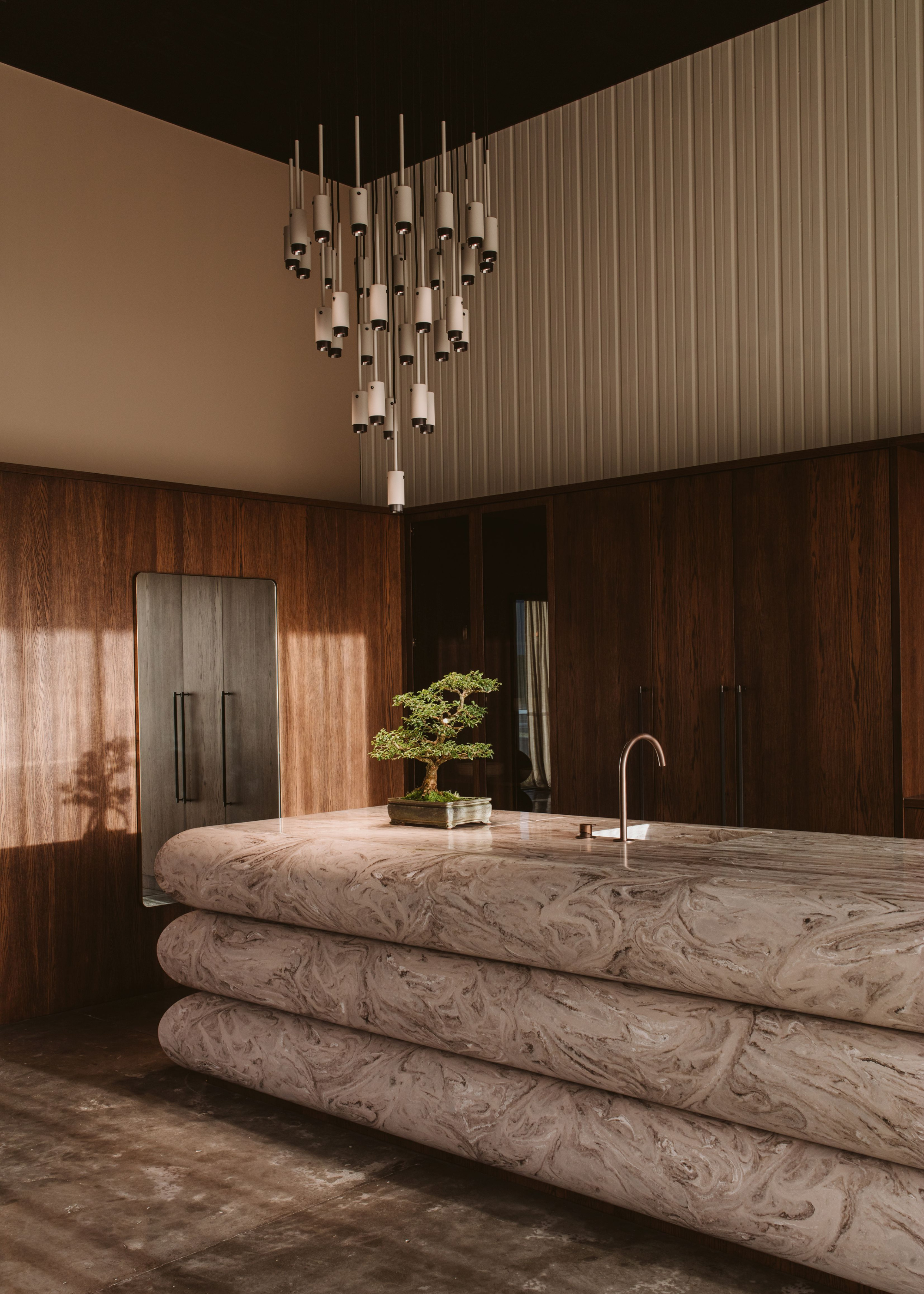 A kitchen with a stacked stone island, a potted bonsai and a mod chandelier