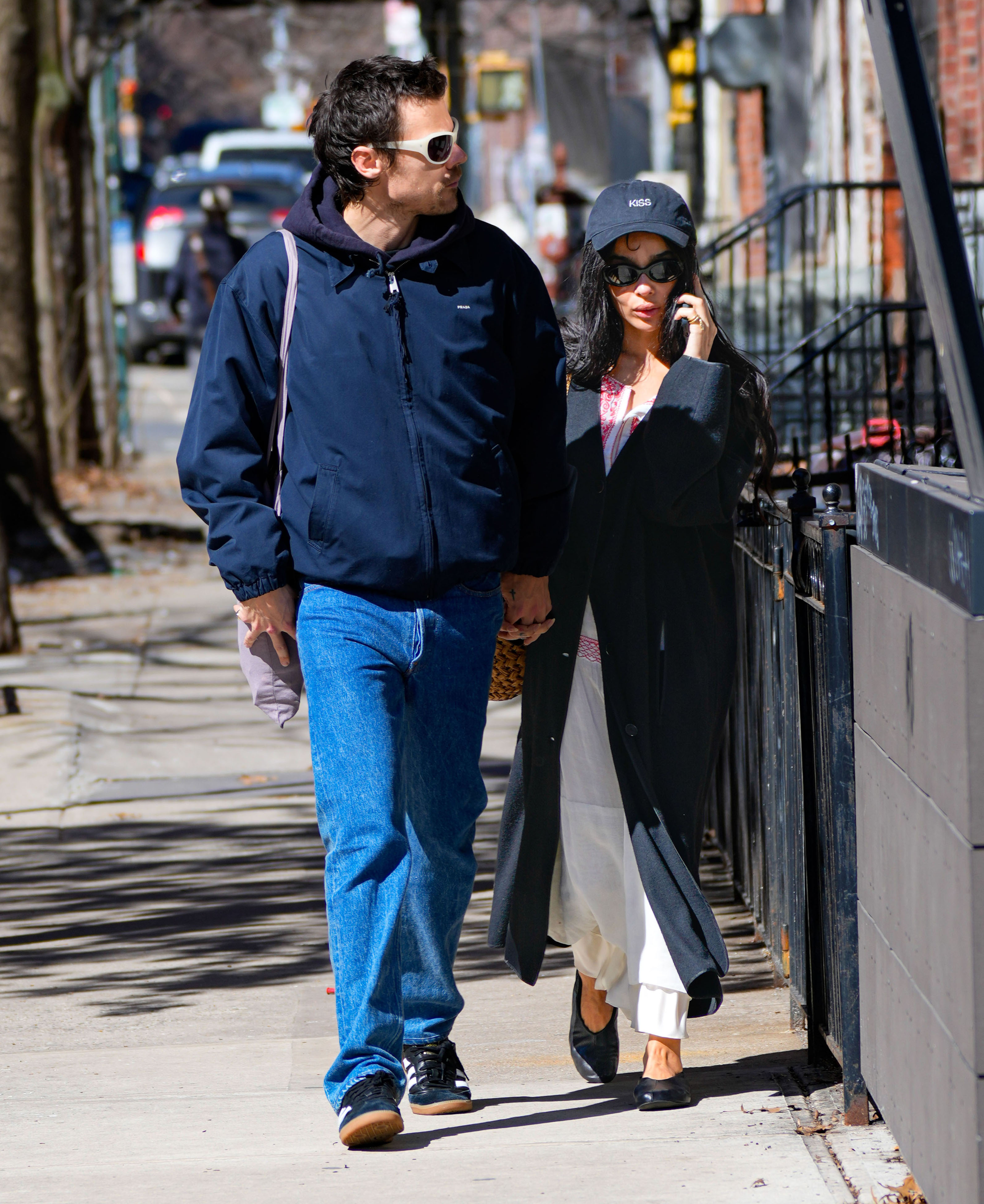 Harry style and zoe kravitz walk in NYC holding hands. Zoe wears a baseball cap, long black jacket, white dress, and black ballet flats.