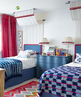 Twin bedroom with two beds with blue throws, canopies, red curtains, blue bed-side table, two table lamps