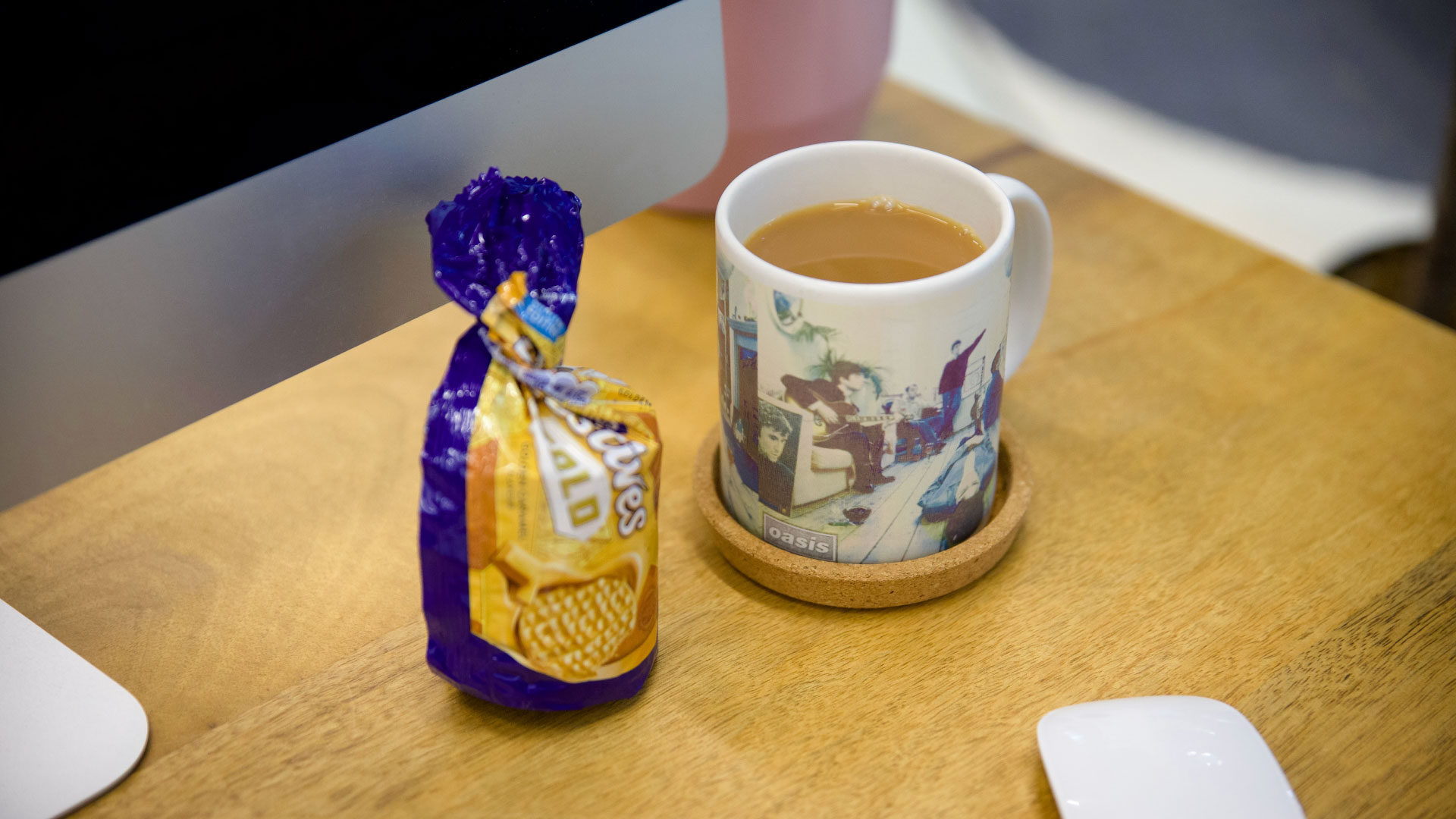 A white mug with a color graphic on it, sitting on a cork coaster, is positioned next to a tightly sealed purple bag of biscuits on a wooden desk.